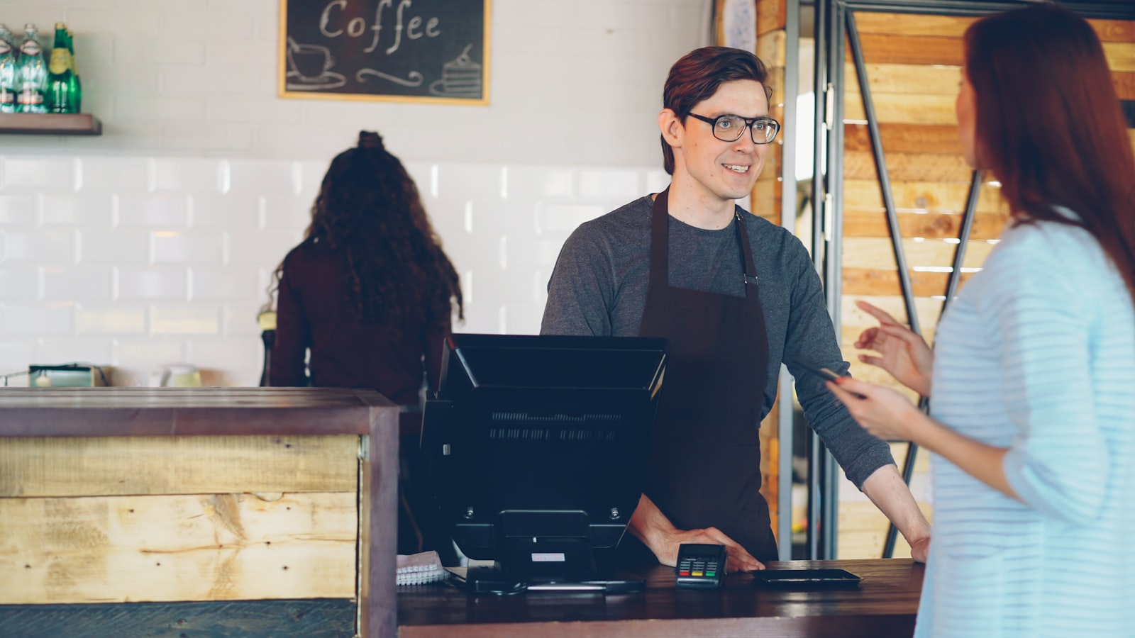 Jeune client réglant sa commande au comptoir d'un café, téléphone en main, le moment idéal pour l'inviter à laisser un avis Google.